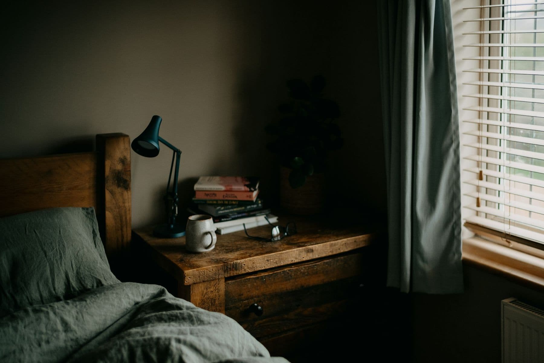 A calm bedroom with a wooden bedside table, a lamp and books beside a window.